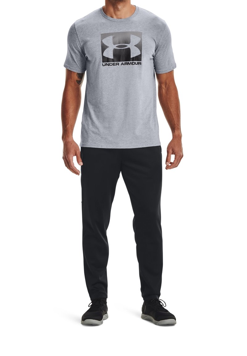 Šedé tričko s krátkými rukávy a velkým černým logem Under Armour. Kombinováno s černými sportovními kalhotami a černými botami, které mají texturovanou tkaninu.