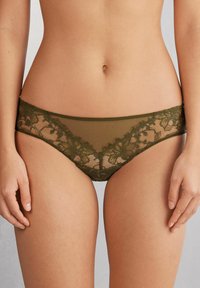 Olijfgroene kanten string met een bloemenpatroon, doorzichtige meshpanelen en een stevige stof. Kenmerken zijn zachte randen en ingewikkelde kantdetails.