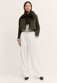 Eine Frau steht vor einem neutralen Hintergrund und trägt eine olivgrüne, kurz geschnittene Jacke, weiße, weit geschnittene Hosen, ein gestreiftes Hemd und einen dunklen Schal.