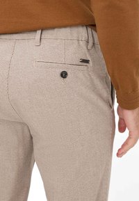 Ternet beige bukser med en baglomme med en sort knap og et lille accentmærke nær taljen. Materialet ser blødt og tekstureret ud.