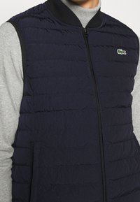 Tmavě modrá péřová vesta se zipem vpředu, žebrovaným límcem a vodorovnou prošívanou strukturou. Má malou zelenou značku a boční kapsy.