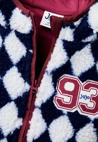 Villāna jaka ar tumši zilu un baltu dimanta rakstu, ar bordo krāsas rāvējslēdzēju un izšūtu "93" ar "JoJo" detaļu.