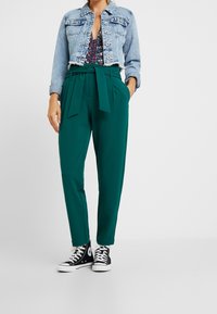 Jeansowa kurtka o kroju cropped, zapinana na guziki, z kwiecistym body pod spodem; zielone spodnie z wysokim stanem, z wiązanym paskiem; czarne sneakersy.