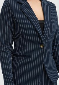 Blazer rayé marine en tissu texturé, présentant des rayures verticales blanches, une fermeture à un bouton et des revers crantés.