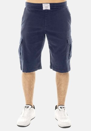 Man wearing navy blue cargo shorts with side pockets and white sneakers, standing against a plain background.