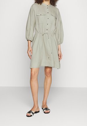 Femme portant une robe vert clair à boutons sur le devant, taille cintrée et manches trois-quarts, assortie à des sandales plates noires et blanches.