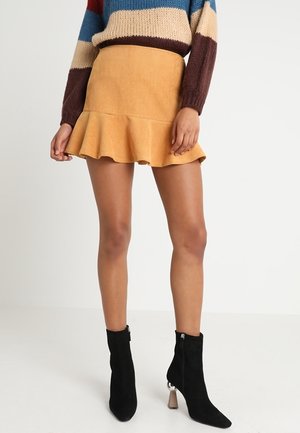 Kvinde iført gul rynket mininederdel, stribet striktrøje og sorte ruskindsankelstøvler med blokhæle, stående mod en ensfarvet baggrund.