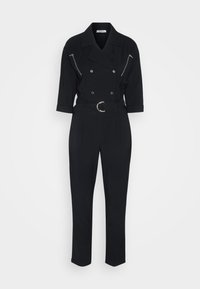 Combinaison noire à manches longues avec boutons croisés, deux poches poitrine zippées, taille cintrée avec ceinture et jambes fuselées.