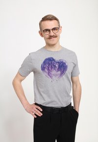 Camiseta de algodón gris con un diseño de corazón morado que muestra caras estilizadas. Combinada con pantalones negros y un cinturón negro.