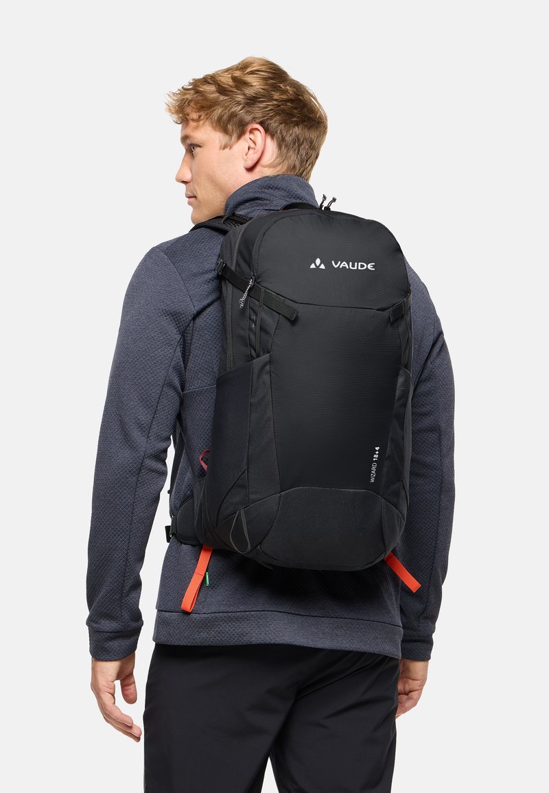 Joven con chaqueta gris oscuro y pantalones negros, llevando una gran mochila negra VAUDE con correas naranjas en la espalda.