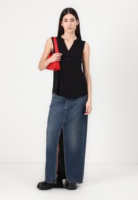 Schwarzes ärmelloses Oberteil mit Knopfleiste, kombiniert mit einem langen Jeansrock mit Schlitz vorne und schwarzen klobigen Schuhen, während eine rote Tasche gehalten wird.