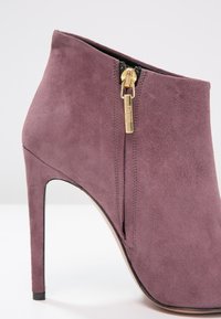 Fioletowe zamszowe botki za kostkę na wysokim obcasie szpilce, z czarnym zamkiem z boku i gładką fakturą. Brak widocznych wzorów.