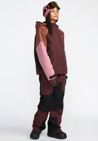 Ensemble d'extérieur d'hiver pour femmes avec un design maron, rose et noir. La veste est dotée d'une fermeture éclair et de poignets ajustables ; le pantalon est ajusté et résistant.