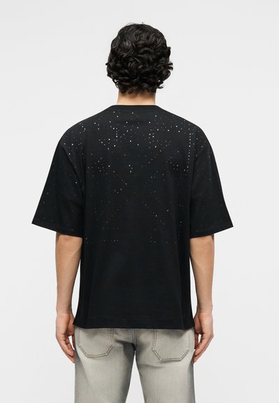 Άνδρας με σγουρά σκούρα μαλλιά που φοράει μαύρο κοντομάνικο πουκάμισο με μικρές οπές και ανοιχτό γκρι τζιν, απαθανατισμένος από πίσω.