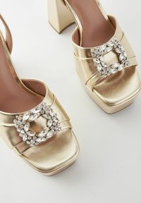 Gold platform sandals with a shiny metallic finish. Features a wide strap adorned with a circular rhinestone detail and chunky heels.