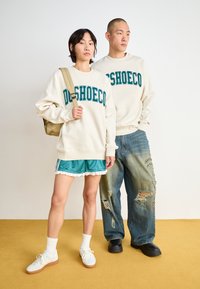 Dva modely stoja na horčicovo sfarbenom povrchu. Jeden má na sebe béžový sveter so teal písaním a teal šortky s čipkovým lemom. Druhý má na sebe béžový sveter a roztrhnuté džínsy. Obe kombinujú s bielymi ponožkami a obuvou.