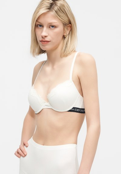 Blond kvinde iført hvid blonder-bh med sort "EMPORIO" bånd, kombineret med hvide højtaljede trusser, stående mod en ensfarvet lys baggrund.