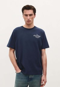 Navy blue cotton t-shirt with short sleeves, crew neckline, and white graphic print on the chest; paired with light blue jeans.