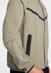 Lätt beige jacka med svarta detaljer, med en framsida som har dragkedja och en sidoficka med dragkedja. Ärmen har en rynkad elastisk manschett.