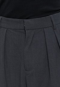 Pantalon plissé gris foncé avec taille et passants de ceinture, porté avec un haut noir rentré.