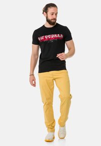 Zwarte katoenen t-shirt met rood en wit grafisch tekstontwerp, gecombineerd met gele broek en witte sneakers.