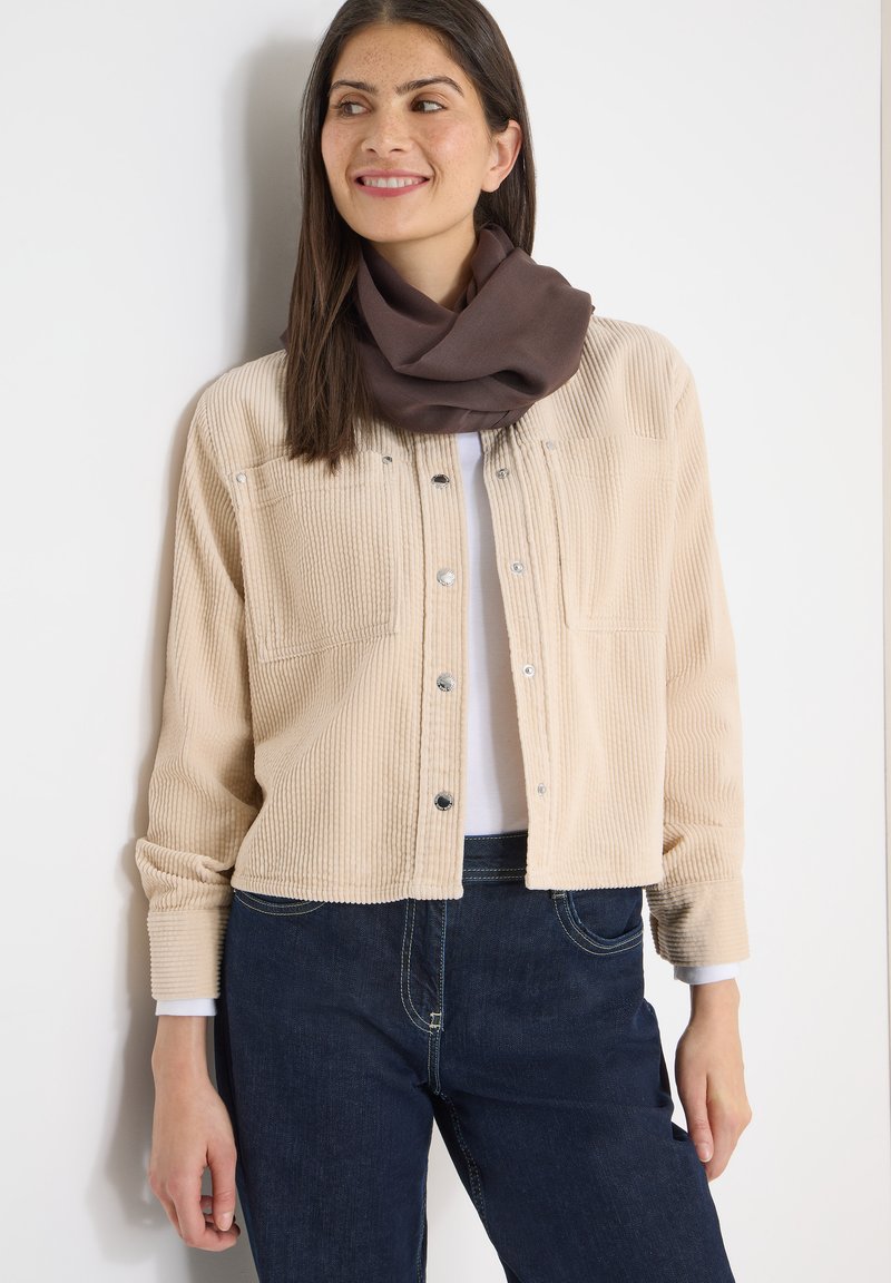 Femme souriante, portant une veste en velours côtelé beige, une écharpe foncée, une chemise blanche et un jean foncé, debout contre un mur blanc.