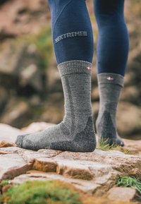 Grå ullsockor med ribbat manschett, strukturerade paneler och förstärkt tå. Har en liten flagga som detalj nära toppen. Fot på stenig terräng.