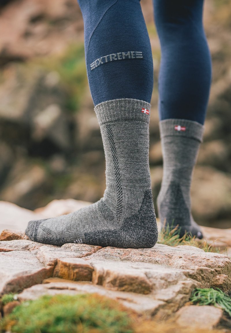 Grå ullsockor med ribbat manschett, strukturerade paneler och förstärkt tå. Har en liten flagga som detalj nära toppen. Fot på stenig terräng.