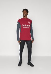 Haut de sport à manches longues rouge et gris avec col zippé, arborant le logo "Emirates Fly Better" ; assorti à un pantalon noir avec des bandes blanches sur les côtés.