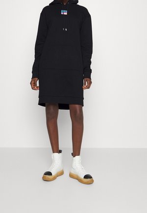 Osoba ubrana w czarną sukienkę z kapturem, z małą kolorową naszywką z logo, oraz białe wysokie trampki z czarnymi noskami i brązowymi podeszwami.