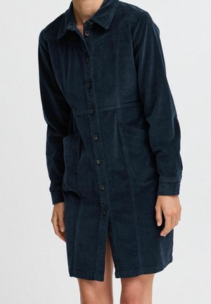 Frau trägt ein langärmeliges, marineblaues Cordkleid mit Knöpfen, Kragen und vorderen Taschen, steht vor einem schlichten hellen Hintergrund.