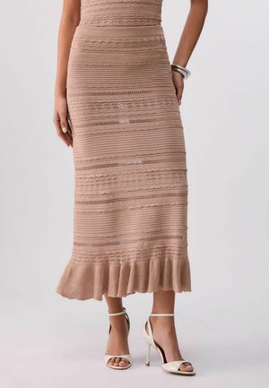 Beige knielanger Strickrock mit horizontalen strukturierten Streifen und gerüschtem Saum, getragen mit weißen Sandalen mit Absatz und Knöchelriemen.