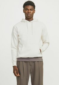 Haut à capuche blanc avec une poche kangourou, un cordon de serrage pour la capuche et des poignets côtelés. Porté sur une chemise gris foncé et associé à un pantalon marron.