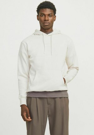 White hoodie with a kangaroo pocket, drawstring hood, and ribbed cuffs. Worn over a dark gray shirt and paired with brown trousers.
