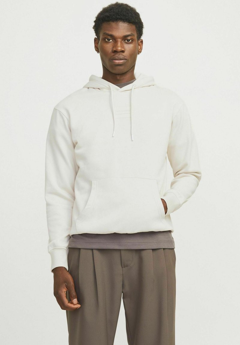 Haut à capuche blanc avec une poche kangourou, un cordon de serrage pour la capuche et des poignets côtelés. Porté sur une chemise gris foncé et associé à un pantalon marron.
