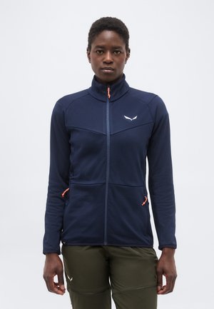 Γυναίκα με μπλε σκούρο μπουφάν με φερμουάρ και ψηλό γιακά και ελιάς πράσινο παντελόνι, στέκεται μπροστά σε απλό φωτεινό φόντο.