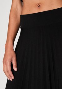 Gros plan sur le torse d’une personne montrant la peau nue et une jupe plissée noire taille haute avec une large ceinture.