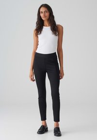 Zwarte slim-fit broek met een elastische tailleband, gecombineerd met een witte mouwloze top. Model staat rechtop met een rechte houding.