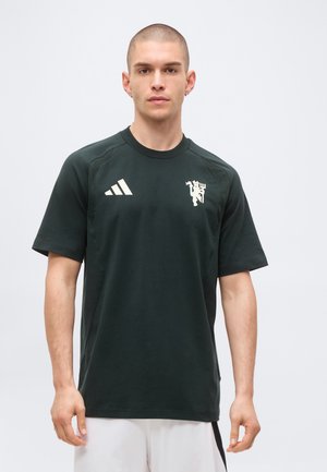 Hombre con cabello corto que lleva una camiseta negra de Manchester United Adidas y pantalones cortos blancos, de pie frente a un fondo claro y sencillo.