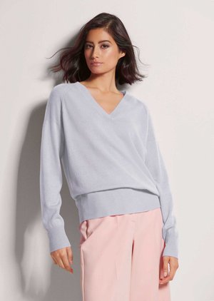 Frau mit schulterlangem dunklem Haar trägt einen hellblauen V-Neck-Pullover und pinke, weite Hosen und steht vor einer hellen Wand.