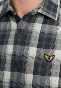 Geruite shirt in marineblauw, grijs en crème. Voorzien van knoopsluiting en geborduurd embleem met vleugels op de borst. Gemaakt van geweven stof.