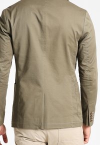 Kurtka w kolorze oliwkowej zieleni wykonana z tkaniny o gładkiej fakturze, charakteryzująca się dopasowanym krojem, rozporkami z tyłu oraz czterema guziczkami zdobiącymi mankiet rękawa.