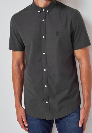 Homme portant une chemise à manches courtes gris foncé boutonnée avec un petit logo brodé sur la poitrine et un jean bleu.