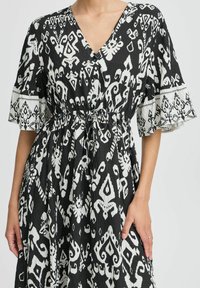 Schwarz-weißes Kleid mit V-Ausschnitt, Kordelzug in der Taille und weit ausgestellten Ärmeln, die mit komplexen Tribal-Mustern und strukturiertem Stoff versehen sind.
