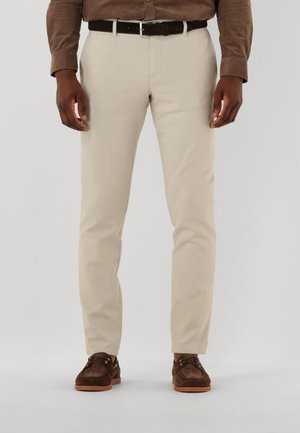 Beige Baumwollhosen mit slim-fit, ausgestattet mit einem Reißverschluss und Gürtelschlaufen, kombiniert mit braunen Wildlederschuhen und einem schwarzen Gürtel.