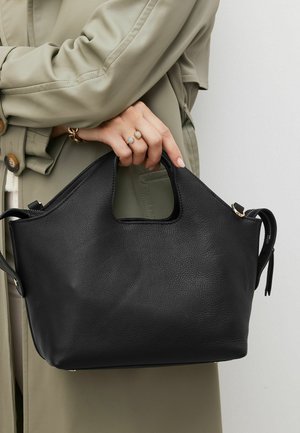 Personne portant un trench-coat beige tient un sac à main texturé noir avec une poignée découpée et une bandoulière.