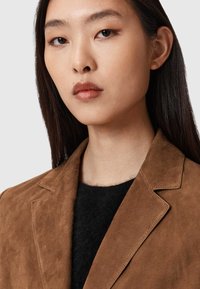 Bruine suède blazer met scherpe revers, textuurafwerking, gedragen over een zwart gebreid topje, waardoor een nette, aangesloten silhouette wordt getoond.