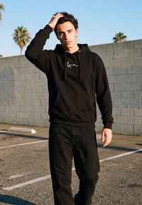 Giovane uomo con felpa nera con cappuccio e pantaloni, in piedi su un'area lastricata accanto a un muro grigio di blocchi di cemento, con palme sullo sfondo.