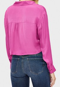 Camicetta corta a maniche lunghe in satin rosa brillante con colletto, dotata di spalle e polsini con bottoni, abbinata a jeans blu scuro.