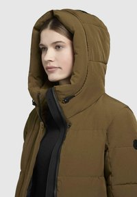 Brown puffer jacket with a large, padded hood. Features a black zipper with adjustable string and a logo patch on the sleeve.